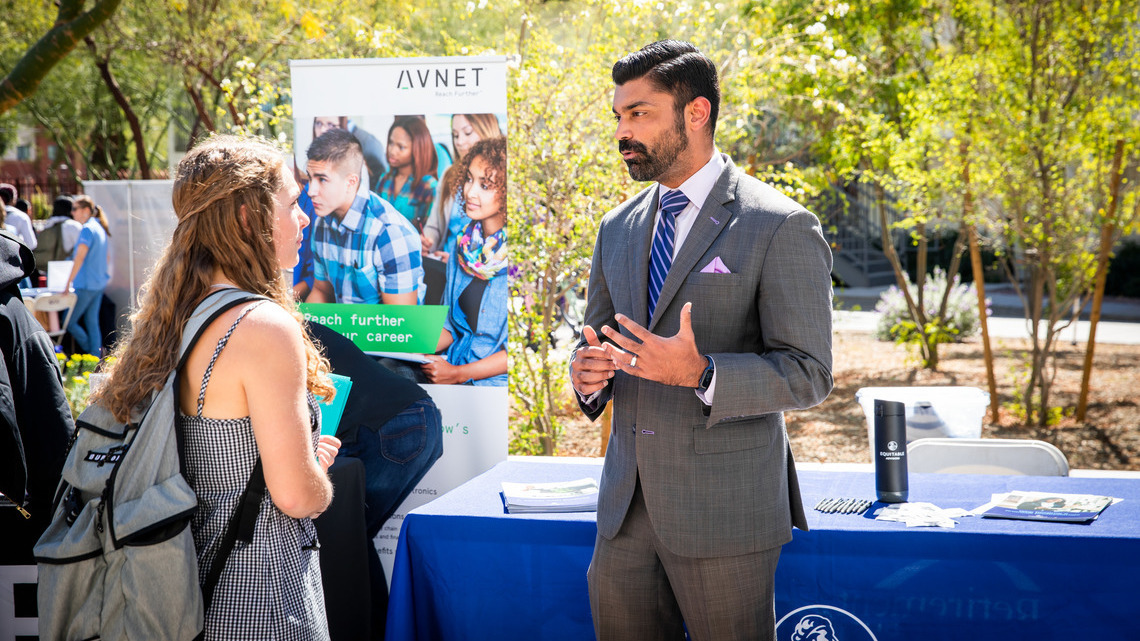 Male recruiter in a gray suit talks to a female student wearing a heavy backpack during an outdoor hiring event at GCU campus.