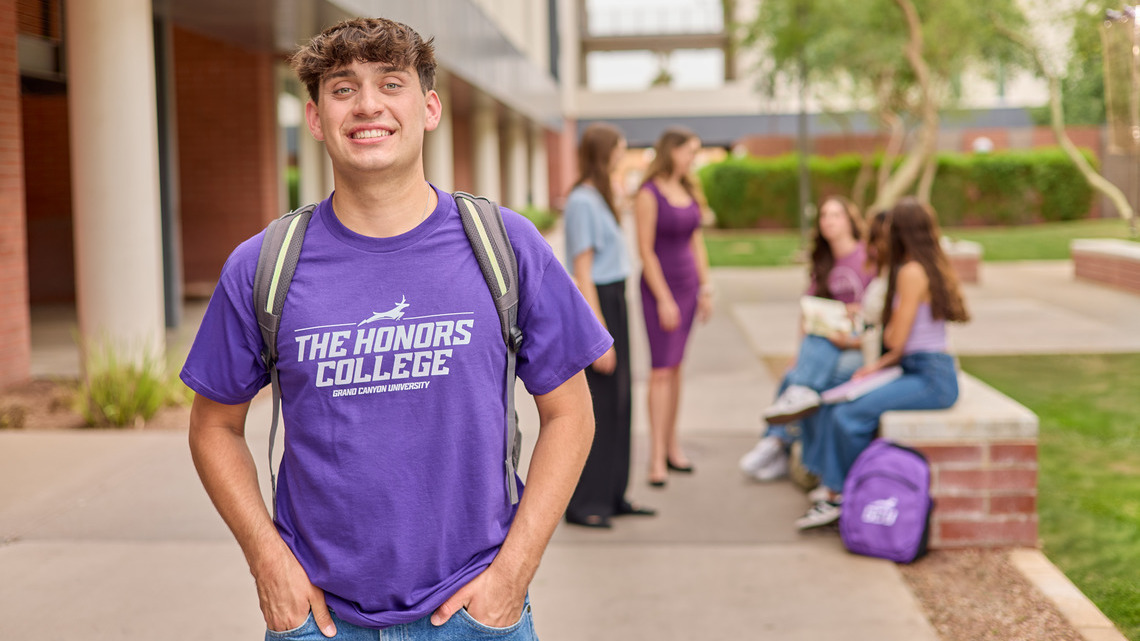 A guy in a purple GCU Honors College-branded shirt stands outside on campus with people behind him.
