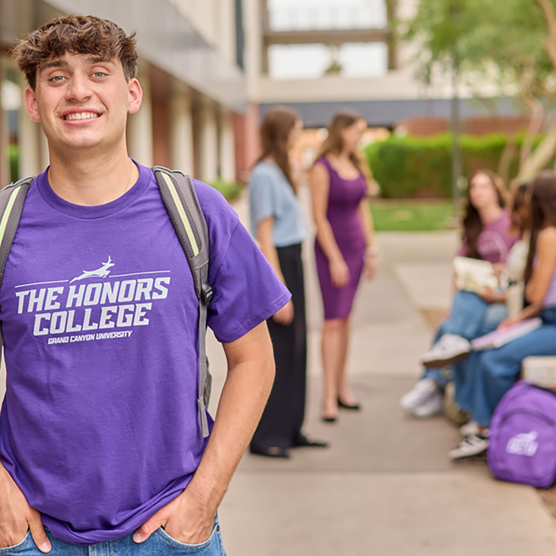 A guy in a purple GCU Honors College-branded shirt stands outside on campus with people behind him.