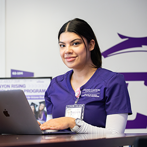 Hispanic female nursing student in purple scrubs with a white long-sleeved undershirt uses laptop with a slight smile.
