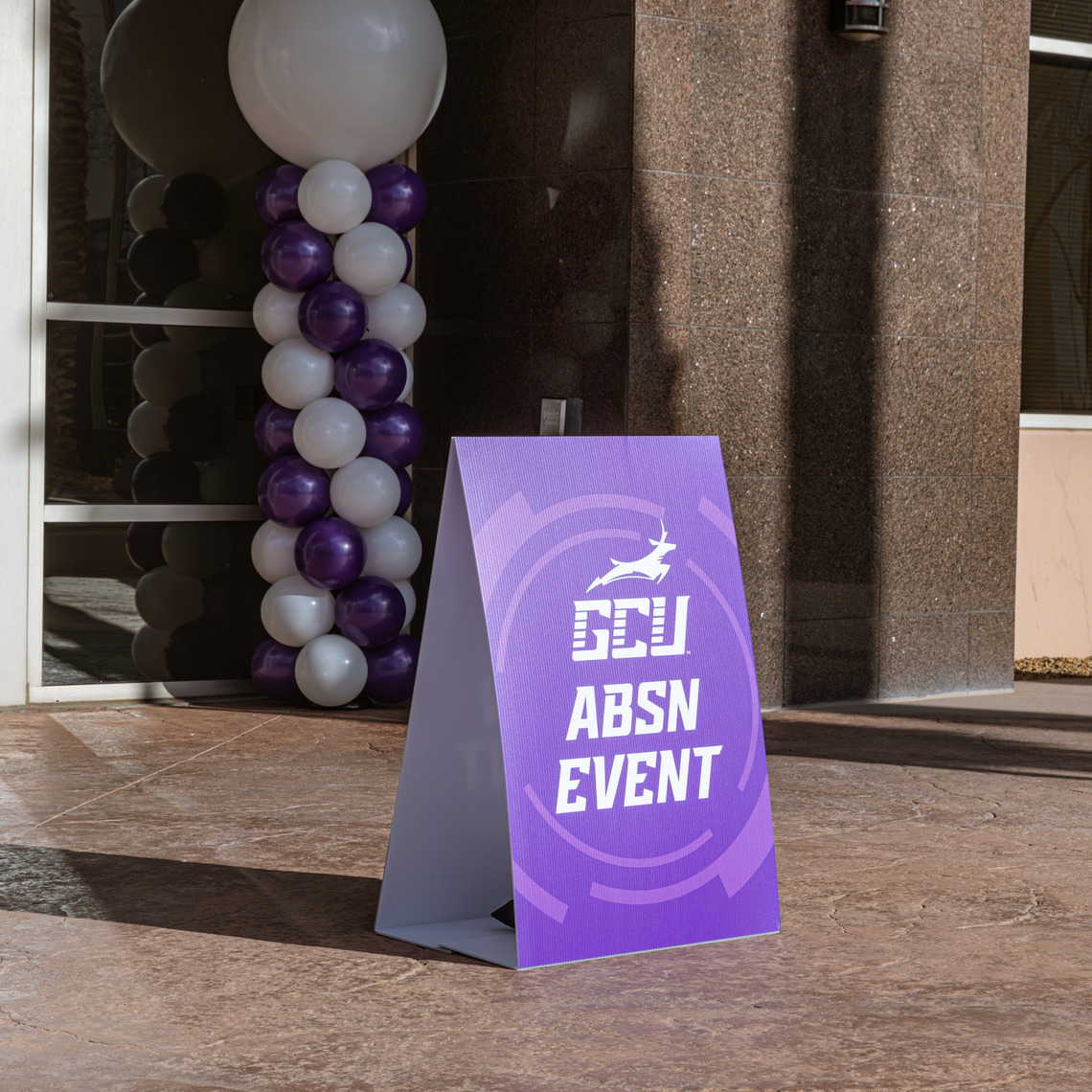 Purple graphic GCU ABSN event a-frame with white and purple balloons set up behind it at the door entrance of an ABSN location launch.