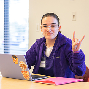 Brunette girl with clear frame glasses gives a GCU lopes up hand sign when using an open laptop while wearing a purple jacket over her GCU ABSN nursing scrubs.