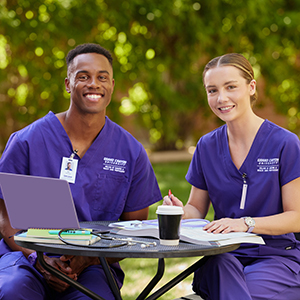 Smiling male African-American and female caucasian nursing students in purple scrubs sitting at an outdoor table to study together.